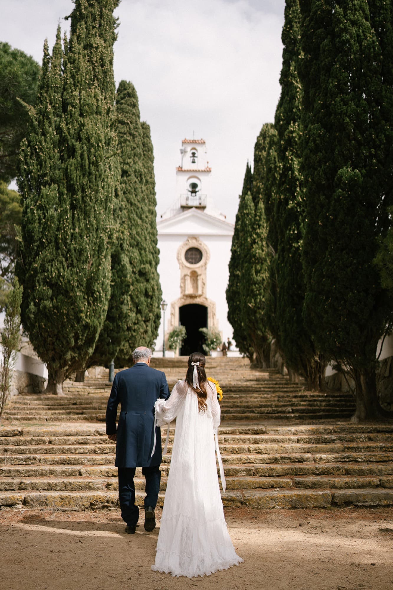 A bride climbing between cypresses towards the church on her wedding day