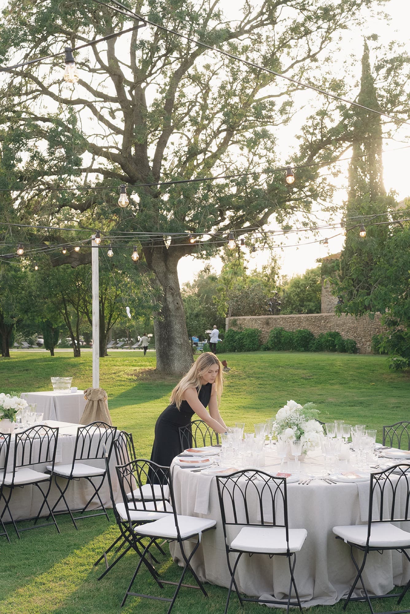 Elisenda Herrero setting a round table in the garden at golden hour, under festoon lights
