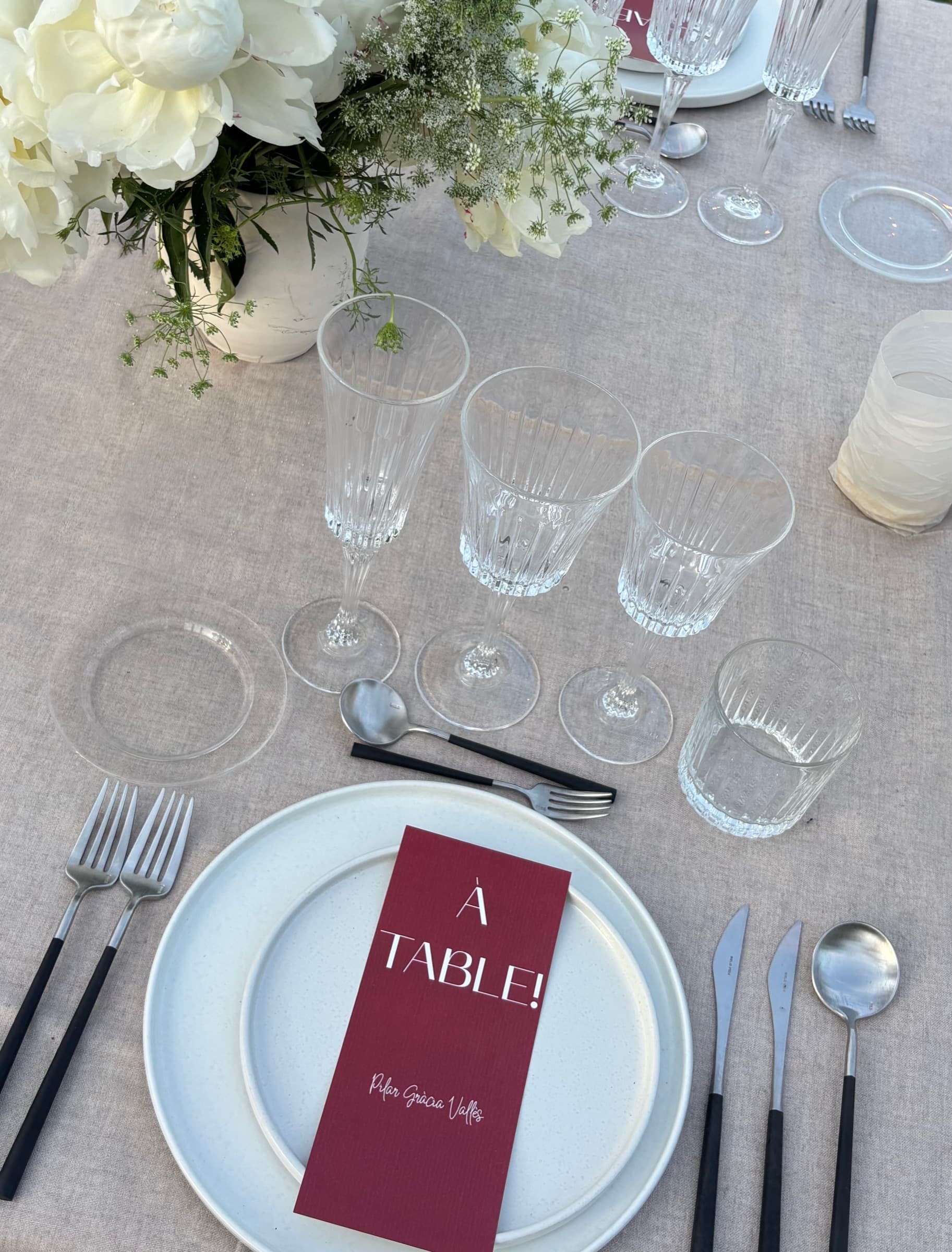 Detalle de una mesa de banquete vista desde arriba: peonías blancas, cristalería tallada y una minuta granate sobre el plato